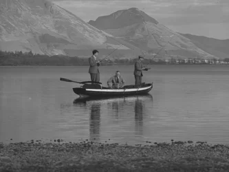 Tre mænd i en robåd på en sø, der fisker, den ene mand er på havet; havlandskab; ted, fjelde i baggrunden