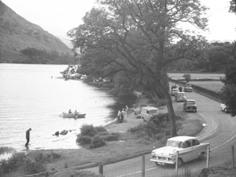 Udsigt over søbredden med mennesker, der står, går, vader og sejler, vejen løber til højre for billedet med parkerede biler