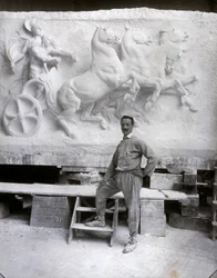 Portræt i fuld længde af den berømte billedhugger Pietro Canonica. Han poserer i sit studie foran et marmorhøjrelief, han arbejder på (s/h-foto)