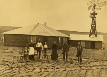Familie af nybyggere uden for deres tørvehus med en vindmølle på taget af den tilstødende bygning i Custer County, Nebraska, ca. 1885