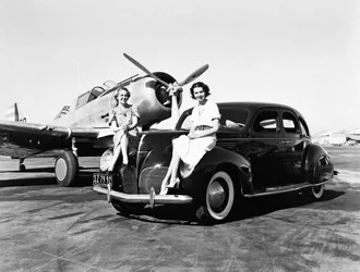 Californien: 1939 To kvinder sidder på skærmene af en Lincoln Zephyr sedan 1939 i en lufthavn med et enkeltmotors fly parkeret bag sig (s/h foto)
