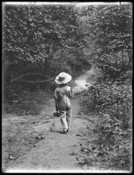William Gray Hassler i stråhat og overalls, bærende fiskestang og kurv, gående ned ad en sti i skoven alene, 20. august 1911