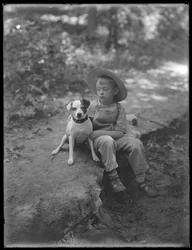 William Grey Hassler i stråhat og overalls, poserede siddende i skoven med en lille hund (Bessie), 20. august 1911