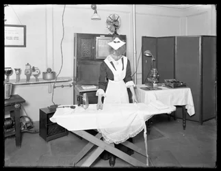 Ung kvinde i stuepigekostume, der demonstrerer brugen af et elektrisk strygejern, ca. 1913-14